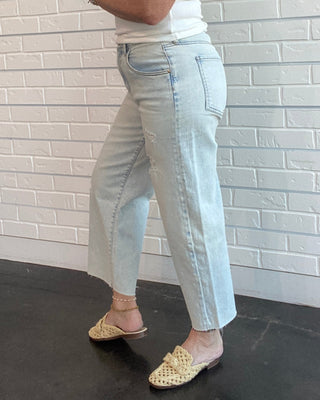 Woman standing against a white brick wall wearing a white tank top and light blue jeans.
