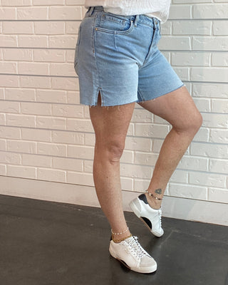 Woman wearing a white top and blue shorts standing against a white brick wall.