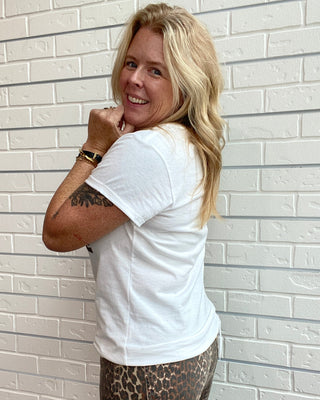 A woman with blonde hair, wearing a white t-shirt and leopard print pants, posing against a white brick wall.