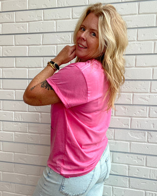 A woman with blonde hair lies on her side against a white brick wall, wearing a bright pink short-sleeve t-shirt and light wash jeans.