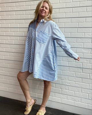 A woman wearing an oversized light blue and white striped shirt dress with a large patch pocket, posing against a white brick wall.