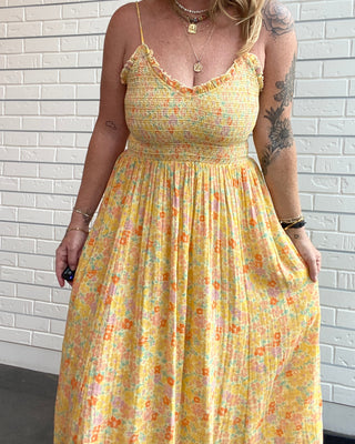 A woman wearing a yellow sleeveless maxi dress with a small multicolor floral print, standing against a white brick wall.