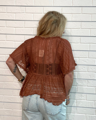 A woman viewed from behind wearing a rust-brown sheer lace short-sleeve peplum top and light-wash jeans, standing against a white brick wall.