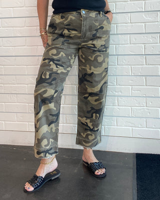 A woman wearing camouflage cargo pants, a black sleeveless top, and black slide sandals, standing against a white brick wall.