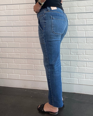 A person wearing blue denim jeans and a black sleeveless top, standing against a white brick-patterned wall.