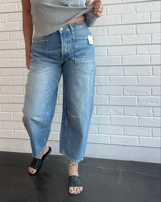 A woman modeling a light gray v-neck t-shirt and wide-leg light-wash blue jeans, standing against a white brick wall.