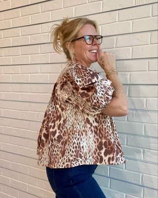 Woman wearing a leopard print top and blue jeans standing against a beige brick wall.