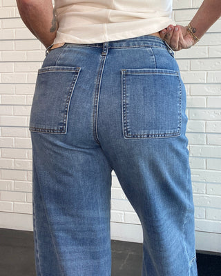 A person wearing light blue wide-leg denim jeans with a cream sleeveless top, shown from the back against a white brick wall.