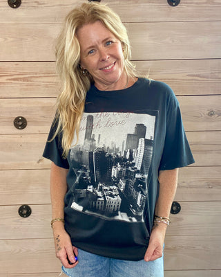 Woman wearing a grey blue t-shirt with a cityscape graphic against a wooden wall.