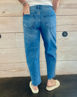 Person wearing blue jeans and a white shirt standing against a wooden wall.