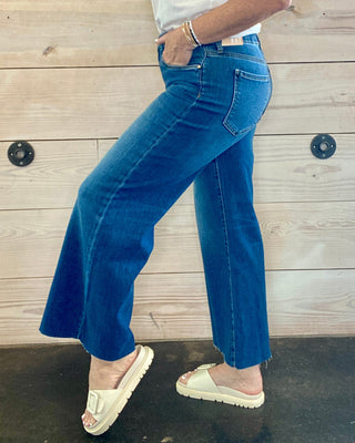 Person wearing a white t-shirt and blue jeans standing against a wooden wall.