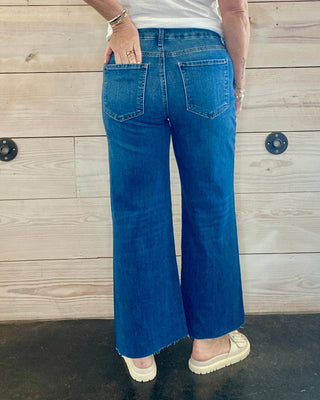 Person wearing a white t-shirt and blue jeans standing against a wooden wall.