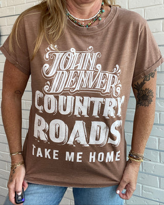 A woman wearing a brown graphic t-shirt with the text 'John Denver Country Roads Take Me Home' in white, paired with blue jeans, standing against a white brick wall.