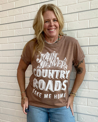 A smiling blonde woman stands against a white brick wall wearing a brown graphic t-shirt with white text that reads 'JOHN Denver COUNTRY ROADS TAKE ME HOME' and blue jeans.
