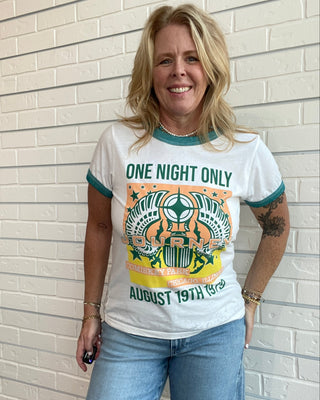 A woman wearing a vintage-style white ringer t-shirt with teal and yellow graphic print that reads 'ONE NIGHT ONLY AUGUST 19TH 1992', paired with light blue jeans, standing against a white brick wall.