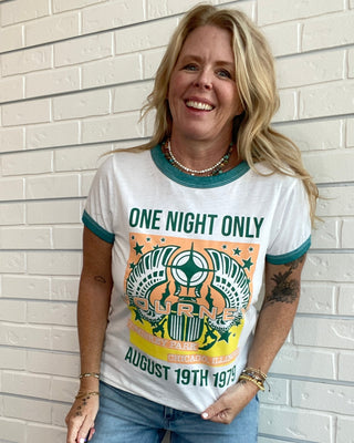 A smiling blonde woman stands against a white brick wall wearing a white ringer T-shirt with teal trim and a large multicolor graphic print, paired with light blue jeans.