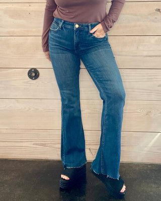 Woman wearing a brown long-sleeve top and blue jeans standing against a wooden wall.