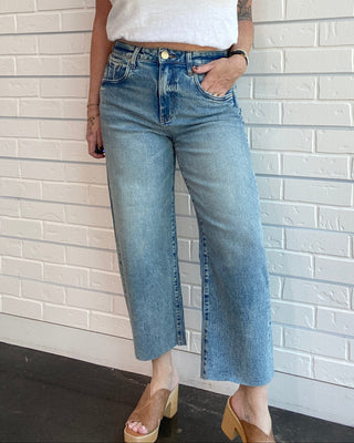 Person wearing a white crop top and blue jeans standing against a light brick wall.