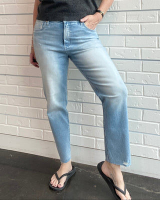 Woman wearing a dark gray sleeveless polo shirt and light blue jeans standing against a white brick wall.