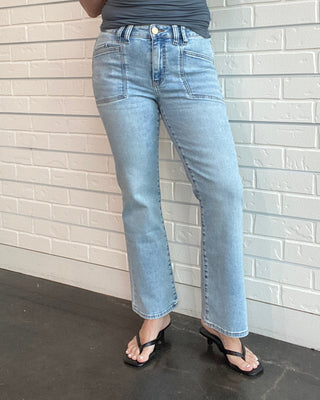 Woman wearing a gray tank top and light blue jeans standing against a white brick wall.