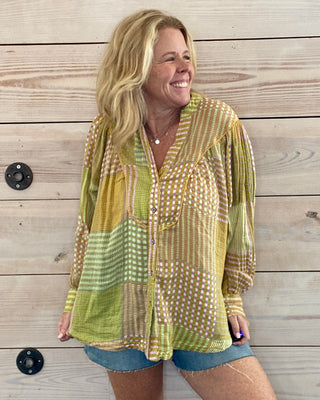 Woman wearing a patterned yellow and green shirt and denim shorts standing against a wooden wall.