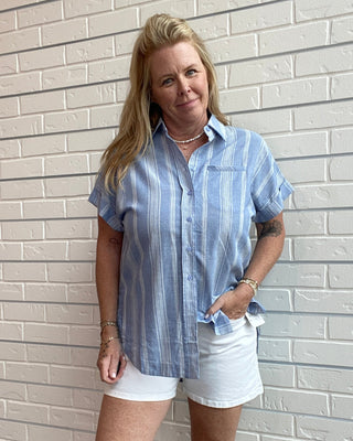 Woman wearing a blue striped shirt and white shorts standing against a light brick wall.