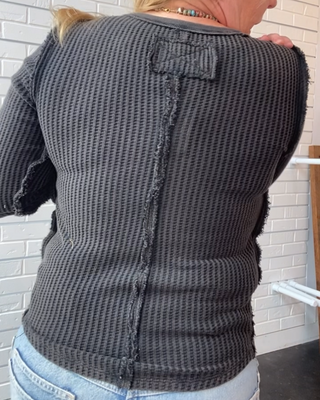 A woman viewed from behind wearing a dark gray waffle-knit cropped sweater with a unique back detail and light blue jeans, standing in front of a white brick wall.