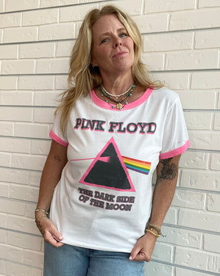 A woman wearing a white ringer T-shirt with pink trim and a Pink Floyd graphic, paired with light blue jeans, standing against a white brick wall.