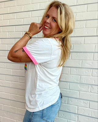 A woman with blonde hair wearing a white ringer t-shirt with pink trim and blue jeans, posing against a white brick wall.