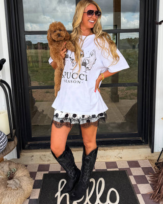 A woman stands on a porch wearing a white graphic T-shirt with 'GUCCI' and 'SEASON.' printed on the front, a black-and-white patterned skirt, and black cowboy boots, while holding a small brown dog.
