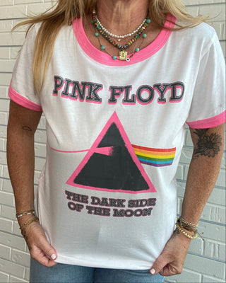 A woman wearing a white ringer T-shirt with pink trim and a large Pink Floyd graphic on the front, standing against a white brick wall.