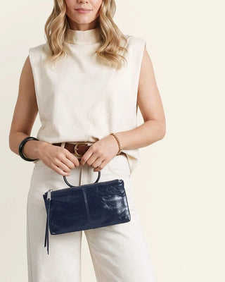 A woman wearing a cream sleeveless mock-neck top and white pants holds a navy blue glossy rectangular handbag with a top handle and tassel detail against a light beige background.