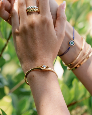 A close-up lifestyle shot of a person's hands and wrists wearing multiple gold-toned jewelry pieces, including a snake-shaped bangle with a small blue stone, a woven ring, and stacked bangles, set against a soft-focus green outdoor background.