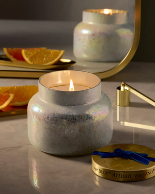 A high blue artisan volcano jar candle with a gold lid, blue ribbon, and iridescent glittery wax inside a clear glass jar,  with a mirror and orange slices in the background