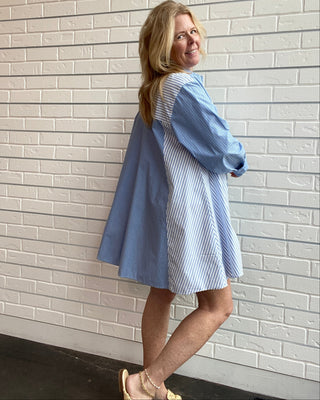 A woman wearing a light blue and white striped shirt dress, posing against a white brick wall.