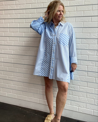 A woman wearing a light blue and white vertically striped button-down shirt dress, standing against a white painted brick wall.