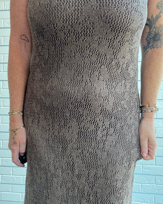 A sleeveless, textured knit maxi dress in taupe brown, shown on a model from the shoulders down against a white brick wall.