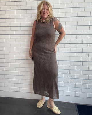 A woman with blonde hair stands against a white brick wall wearing a sleeveless, ribbed brown knit midi dress and beige woven mule sandals.
