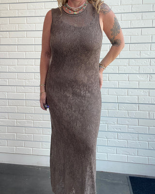 A woman wearing a sleeveless, knee-length brown knit maxi dress with a sheer mesh overlay, standing against a white brick wall.