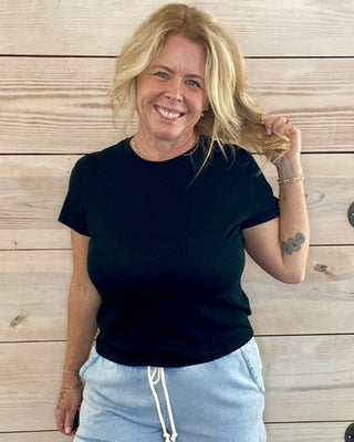Woman wearing a black t-shirt and light blue shorts standing against a wooden wall.