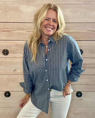 Woman wearing a grey striped shirt and white pants standing against a wooden wall.