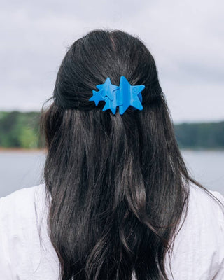 Person with blue hair clips in their dark hair, standing outdoors with a lake and trees in the background.