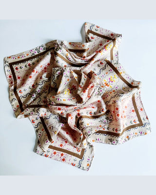 A square scarf with a beige base, decorated with a colorful floral pattern and brown wavy borders, laid out in a loose, draped arrangement on a light gray background.