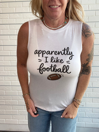 A woman wearing a white sleeveless graphic tank top with the text 'apparently I like football' and a small football graphic, paired with blue jeans, standing against a white brick wall.
