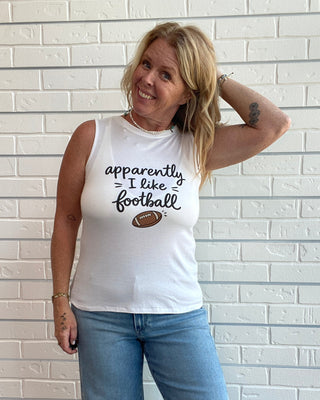 A woman wearing a white sleeveless tank top with the text 'apparently I like football' and a small football graphic, paired with blue jeans, standing against a white brick wall.