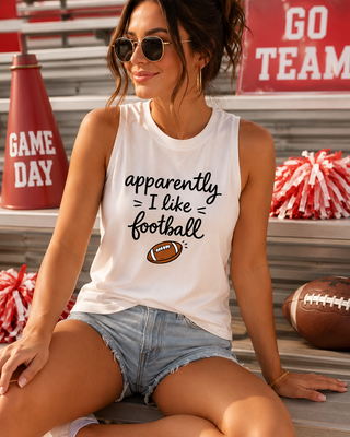A woman sitting on stadium bleachers wearing a white graphic tank top that reads 'apparently I like football' with a small football graphic, denim shorts, and sunglasses. A red cone, pom-poms, and a football are visible around her.