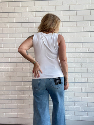 A woman viewed from behind wearing a white sleeveless tank top and blue jeans, standing against a white painted brick wall.