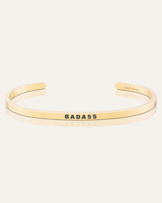 A slim gold-tone cuff bracelet with the word 'BADASS' engraved in black capital letters on the front, shown on a light cream background.