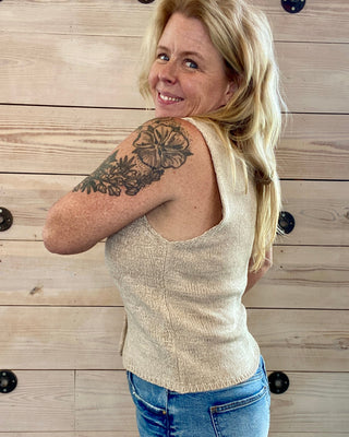 Woman with a tattoo on her arm wearing a beige sleeveless top and blue jeans against a wooden wall.