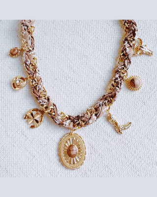 A bohemian-style gold chain necklace with woven brown and beige fabric details, featuring a central oval pendant with a stone, crescent moon charm, and other decorative charms, displayed on a textured off-white fabric background.
